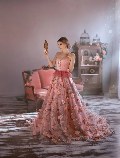 Красивая женщина в сексуальном длинном средневековом розовом платье, весенние цветы на юбке, смотрит на себя. Держит в руках винтажное зеркало. Взрослая королева с большой грудью в корсете, с открытой грудью. Элегантные волосы булочки — стоковое фото
