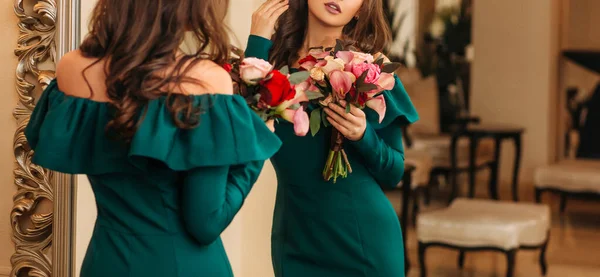 Σιλουέτα της νεαρής όμορφης γυναίκας σε πράσινο μοντέρνο σμαραγδένιο βραδινό φόρεμα, κοιτάζοντας τον εαυτό της στον καθρέφτη, κρατώντας μπουκέτο λουλούδια στα χέρια της. κλασικό πολυτελές αραβικό εσωτερικό. Πρόσωπο κομμένο. Ηνωμένα Αραβικά Εμιράτα — Φωτογραφία Αρχείου