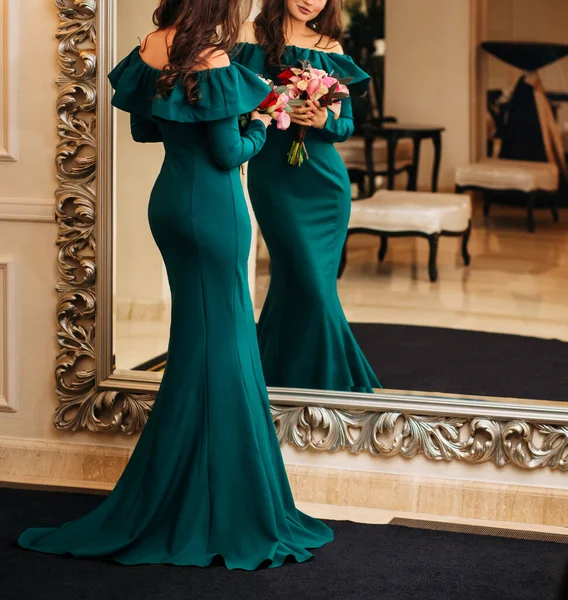 Μυστηριώδης νεαρή όμορφη κομψή γυναίκα σε πράσινο σμαραγδένιο βραδινό φόρεμα, η σιλουέτα μιας γοργόνας στέκεται κοιτάζοντας τον εαυτό της στον καθρέφτη, κρατώντας ένα μπουκέτο λουλούδια στα χέρια της. κλασικό εσωτερικό πολυτέλεια — Φωτογραφία Αρχείου