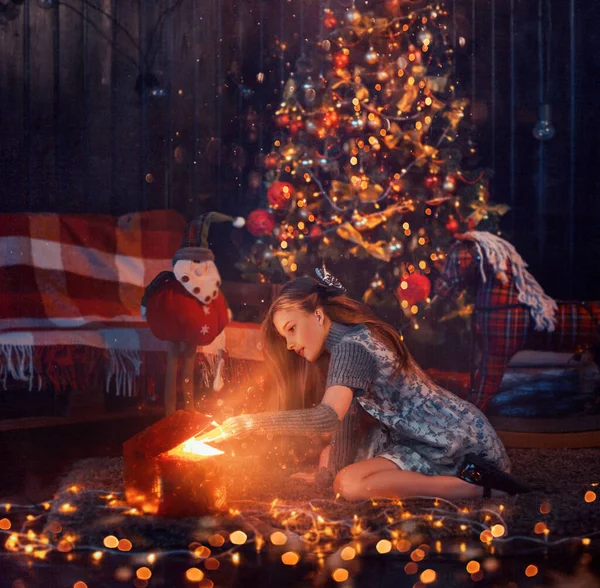 Una niña feliz se sienta en el suelo en una habitación oscura bajo un árbol de Navidad y abre regalos. La luz mágica emite una caja de regalo abierta. Misticismo, magia, milagro en la noche de Navidad. Vacaciones de Año Nuevo —  Fotos de Stock