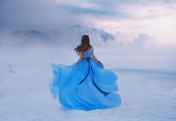 Mistério fantasia Mulher Rainha da Neve em azul vestido exuberante, voar no vento. Senhora viajante. Arte fundo inverno congelado natureza montanhas, neve branco céu dramático nuvens céu. Menina princesa caminha, vista traseira — Fotografia de Stock