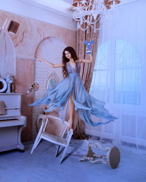 Χαρούμενη χαρούμενη ευτυχισμένη νεαρή γυναίκα πετάει γύρω από το δωμάτιο με ανεμιστήρα στα χέρια της. αστεία φωτογραφία αιώρησης. Ιστορικό λευκό δωμάτιο πολυθρόνα παράθυρο. Μπλε φόρεμα φτερουγίζει στον αέρα. Νεράιδα πνεύμα κορίτσι — Φωτογραφία Αρχείου