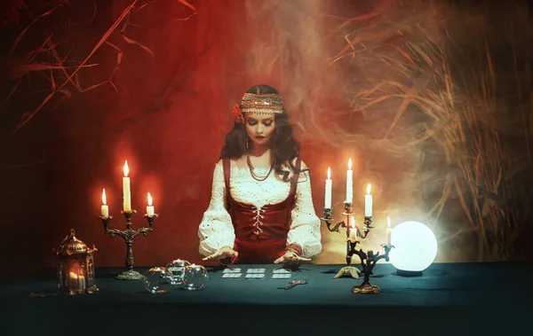 Fantazie dívka v obrazu cikánské čarodějnice sedí u stolu v tmavě gotické místnosti. Kostým Art Red. Kouzelná žena věštkyně čte budoucnost na tarotových kartách, křišťálová koule. Rituální hořící svíčky, seance, kouř — Stock fotografie