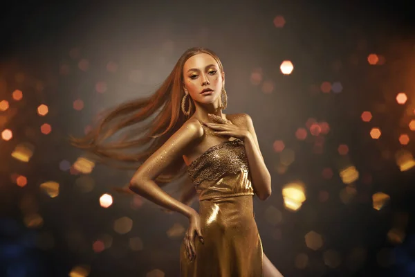 Fiatal elegáns lány divat modell arany ruha, ruhát pózol stúdióban. Hosszú szőke haj repül a szélben. Gyönyörű nő arany bőrrel festve. Háttér világítás, csillogás, sárga bokeh — Stock Fotó