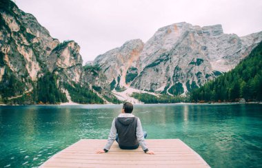 Ellerine yaslanmış bir adam ahşap iskelede oturuyor Braies Gölü 'nde, güzel, gizemli, beyaz dağların tadını çıkarıyor. Koyu kısa saçlı adam. Sahildeki yeşil orman zümrüt su yüzeyi