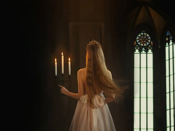 Μυστηριώδης τέχνη μεσαιωνική πριγκίπισσα κορίτσι βόλτες στο σκοτεινό γοτθικό δωμάτιο. Γυναίκα βασίλισσα κρατά κηροπήγιο με αναμμένα κεριά στο χέρι. Φόρεμα με ανοιχτή πλάτη μακριά χαλαρά ξανθά μαλλιά που πετούν σε κίνηση. Φύγε. — Φωτογραφία Αρχείου