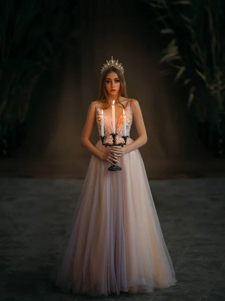 Photo d'art de la princesse médiévale se promène dans la chambre gothique sombre. Femme reine tenant chandelier avec des bougies brûlantes à la main. Robe rose pourpre, longs cheveux blonds lâches, couronne royale dorée. Image de fée — Photo