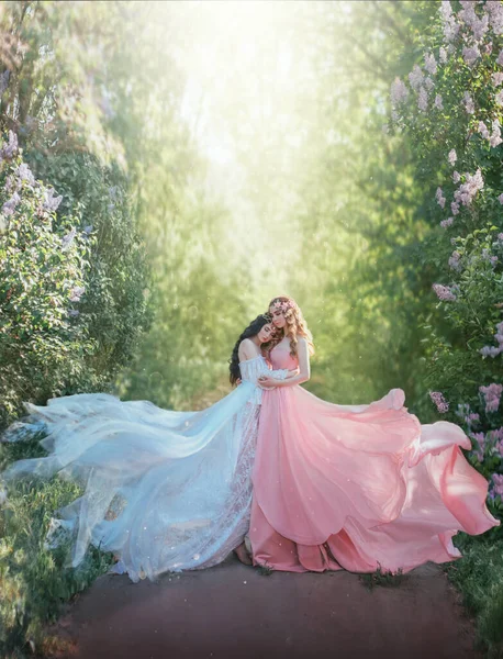 Fantazie fotografie Dvě mladé krásné okouzlující ženy stojí objímají v jarní zahradě na pěšině. Žena princezna v bílém hedvábí vintage šaty tkaniny létání ve větru. Královna v růžových šatech. Zelené přírodní stromy — Stock fotografie