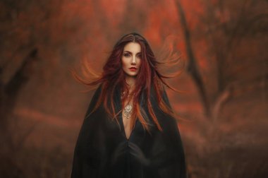 Fantezi gotik kadın kara cadı. Siyah pelerinli kızıl saçlı şeytan kız şeytan. Uzun saçlar rüzgarda uçuşuyor. Koyu yoğun sonbahar ormanı portakal rengi ağaçlar. Ortaçağ elbisesi, ipek elbiseler 