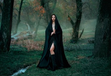 Esrarengiz fantezi gotik kadın. Kötülüğe takıntılı kara cadı. Siyah pelerinli dişi vampir. Karanlık, sık ormanın arka planında yürümek, ağaçlar. Ortaçağ kraliçesi ipek pelerinli, eşarplı