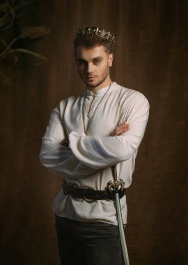 Ortaçağ kralına benzeyen yakışıklı bir adamın portresi. Eski moda kıyafetler, tarihi beyaz gömlek eski moda altın kaplama başlık. Gotik oda, stüdyo. Moda modeli pozu. Yetişkin güçlü seksi adam şövalye.