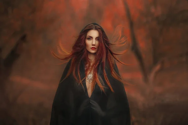 Fantasy gothic woman dark witch. Red-haired evil Girl demon in black dress cape hood. Long hair flutters fly in wind. Dark dense deep autumn forest orange colors trees. Medieval dress, silk clothes — Stock Photo, Image