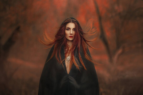 Fantasy gothic woman dark witch. Red-haired evil Girl demon in black dress cape hood. Long hair flutters fly in wind. Dark dense deep autumn forest orange colors trees. Medieval dress, silk clothes.