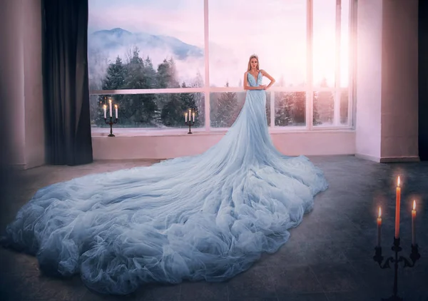 Jovem mulher loira fantasia princesa Cinderela em glamourosa vestido azul fica na janela enorme quarto branco. Saia longa tule bainha trem. Imagem romântica elegante rainha medieval, coroa. Menina senhora histórica — Fotografia de Stock