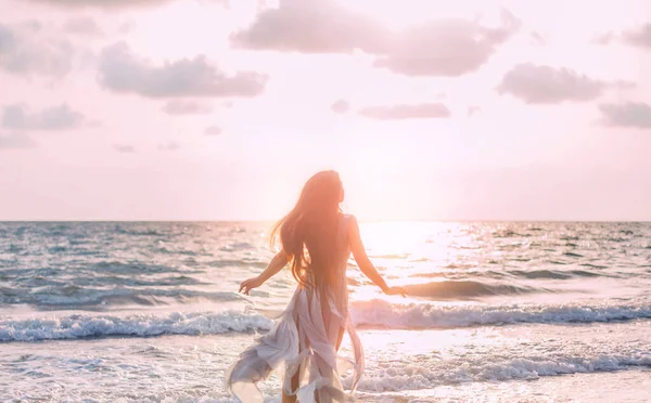 Silhouette de belle jeune femme heureuse promenades touristiques court par le littoral. fille admire la beauté lumière du soleil divin lumineux coucher de soleil de mer. Ciel bleu nuageux rose. Vue arrière des cheveux longs flottants. robe de soie mouche — Photo