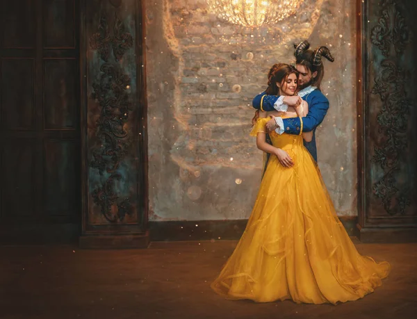 Abbraccio di coppia in camera vecchio castello. Felice bellezza donna principessa fantasia in abito giallo e ragazzo è bestia incantata, corna sulla testa. Romantico principe maschio abbraccia ragazza in armi. Uomo mostro carnevale costume. — Foto Stock