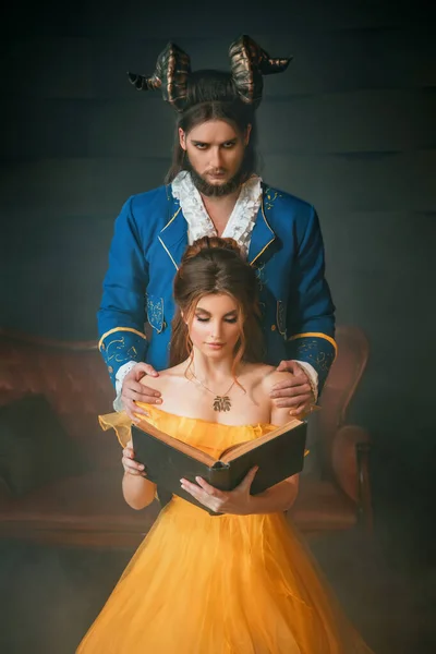 Fantasy man enchanted prince with rams horns on head, hugging a beautiful woman by shoulders. Girl in yellow medieval historical dress holds reads vintage book. Gothic dark room. Guy beast monster — Stock Photo, Image