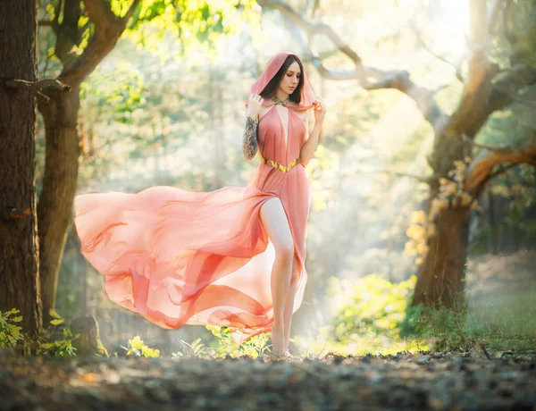 Young beautiful woman fantasy elf walks in spring forest. Green trees background, summer park. Girl princess in orange peach color silk dress, hood cape flies waving in wind motion. Sexy fashion model — Stock Photo, Image