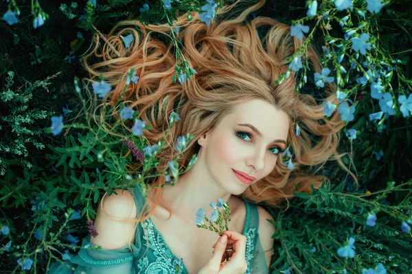 Art photo fairy tale beauty. Portrait Fantasy woman lies on blooming meadow in blue vintage dress. Summer nature background spring green grass flowers. Girl princess blond long hair. Look at camera — Stock Photo, Image