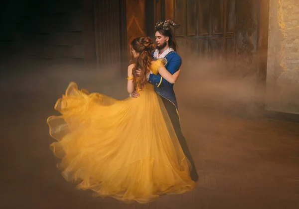 Couple danse au bal fantastique. Joyeux beauté femme princesse en robe jaune et mec bête enchantée, cornes sur la tête Fille tourbillonne dans les bras du prince mâle. Costume monstre homme. Mouche en tissu de soie en mouvement — Photo