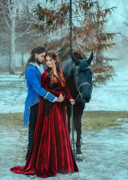 Medieval couple in love man and woman hugging in winter forest. Vintage clothing red long dress. Blue frock coat costume, tailcoat caftan. Prince and princess together. Black steed horse. Art image