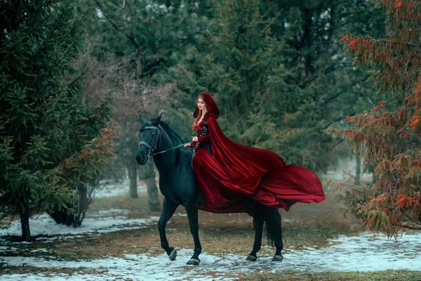 Medieval woman princess in red dress sits astride black steed horse. Girl rider in vintage cloak cape train flies in wind motion. Background green trees spruce forest spring winter nature melted snow — Stock Photo, Image