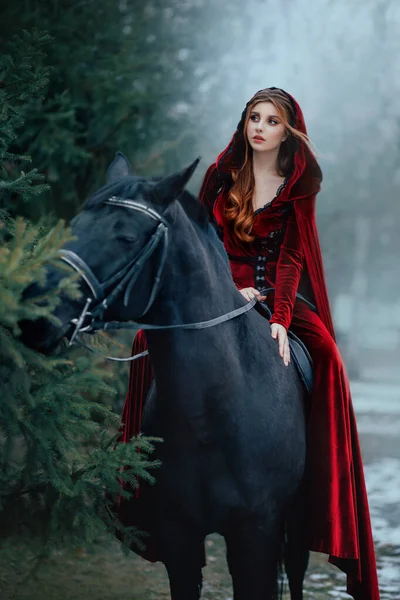 Medieval woman princess in red dress sits astride black steed horse. Girl rider in vintage cloak cape train flies in wind motion. Background green trees spruce forest, spring winter nature melted snow — Stock Photo, Image