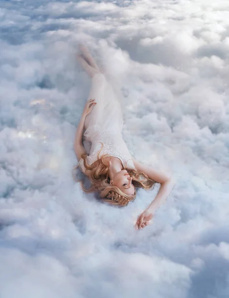 Jonge mooie blonde vrouw ligt in zacht bed op witte wolken in de lucht. Fantasie meisje slapende schoonheid. Godin die droomt van het leven op de top van de hemel Olympus. Goddelijke licht dame engel — Stockfoto