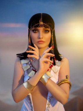 Mısır stili kadın portresi. Seksi tanrıça Kraliçe Kleopatra çöl piramitlerinde duruyor. Sanat tarihi firavun kostümü, beyaz elbise, altın aksesuarlar. Mısır makyajı. Yaratıcı saç bandı, yılan bileziği.