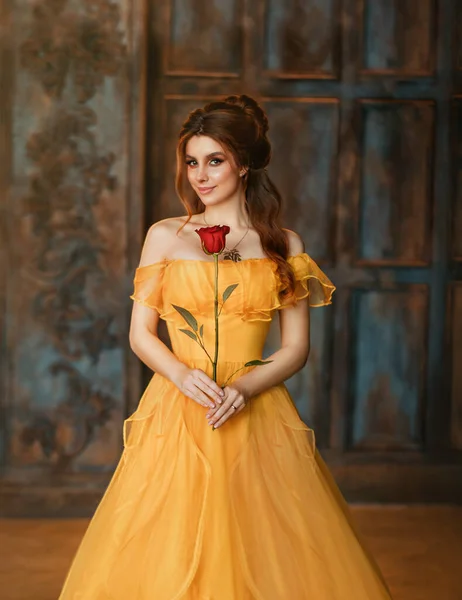 Girl beauty fantasy princess in yellow long historical, medieval silk dress holding flower red rose in her hands. Background of old gothic castle room. Fairy tale bewitched queen. Happy woman smiles — Stock Photo, Image