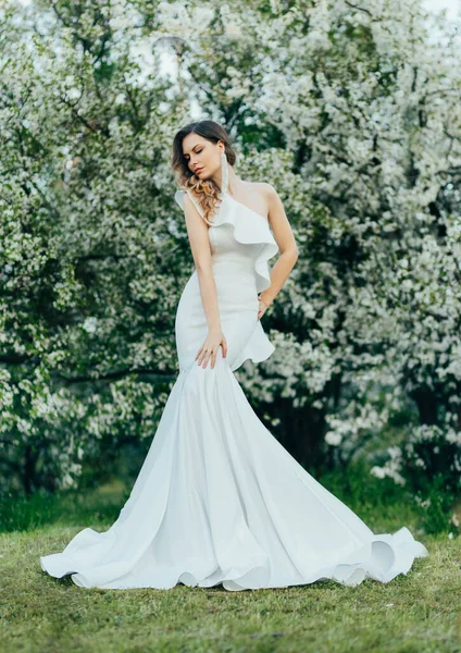 Ung vacker kvinna modell poserar i vit aftonklänning silhuett sjöjungfru. Snygg tjej brud perfekt klänning, långt tåg. Natur bakgrund gröna blommande träd. Vår bröllop körsbär trädgård. — Stockfoto