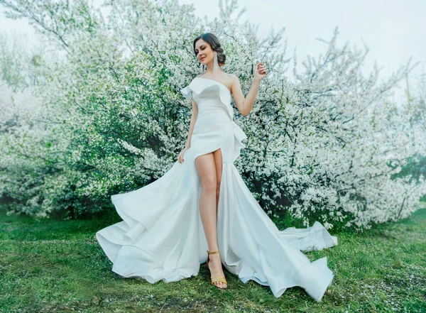 Fantasy nő hosszú fehér elegáns divat hosszú ruha séták zöld tavaszi virágos cseresznye kertben. Boldog vidám lány hercegnő menyasszony szoknya szövet legyek áramló hullámzó szélmozgás. Szexi csupasz lábak — Stock Fotó