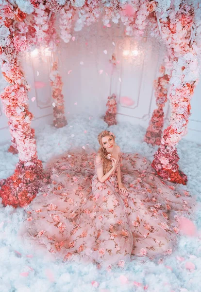 Фантазія весна щаслива жінка в довгій сукні. Тканинна спідниця в квітах. Творча арка червоно-рожевих білих троянд. Пелюстки падають. Казкова принцеса дівчина сидить на зимових снігових пейзажах, кімнаті художнього декору. Модна модель — стокове фото