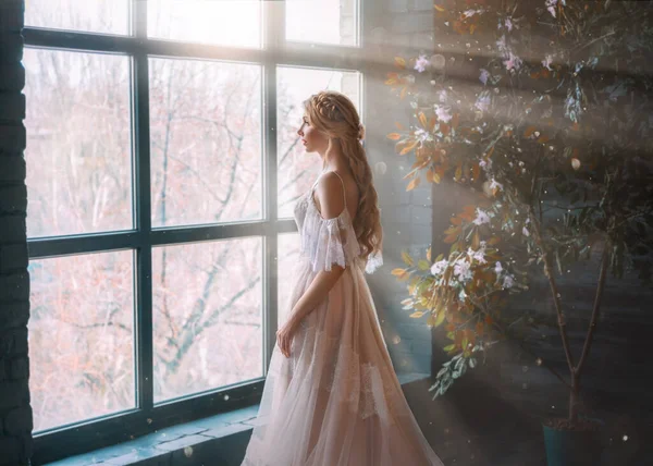 Senhora romântica, mulher loira com cabelos longos em vestido vintage branco fica no quarto escuro, olha para a janela. Menina princesa noiva em vestido de noiva. Penteado elegante. Raios brilhantes de sol conceito de espera — Fotografia de Stock