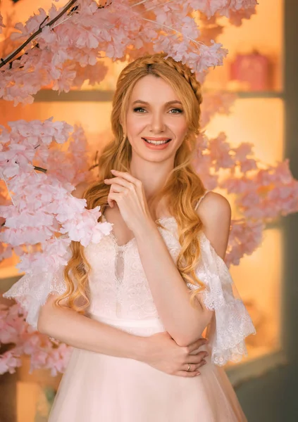 Portrait of young happy beautiful blonde woman in white vintage dress. Girl laughs smile on face. Studio background, blooming pink sakura tree. Long hair is braided into an elegant romantic hairstyle — Stock Photo, Image