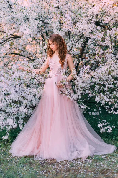 Young red-haired girl princess in pink evening tulle fluffy dress. Elegant romantic woman walking in blooming cherry garden. Enjoyment of Spring Nature. Green trees white flowers. Fantasy vintage lady — Foto Stock