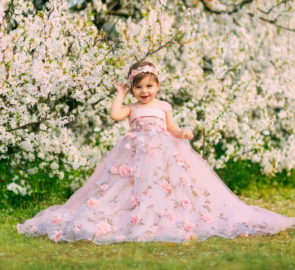 Mladá šťastná malá víla princezna. Šťastná dětská tvářička se usmívá. Luxusní nadýchané dlouhé šaty pro děti, růžové oblečení. Bílé květy pozadí stromy jarní příroda. Módní model, rok starý. — Stock fotografie