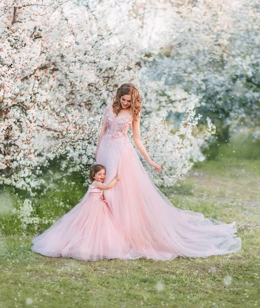 Young happy family, mom and daughter in beautiful evening festive long powdery pink color dresses. Woman holds little girl in hand, looks with love, smile on face. Wedding lush outfits. Nature spring. — Stockfoto
