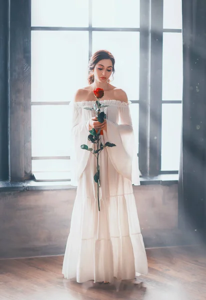 Fantasy vintage beauty woman sexy bare shoulders. white long medieval dress wide sleeves renaissance style. Lady holds red rose in hands stands on backdrop window with magical bright sun ray sunlight. — Zdjęcie stockowe