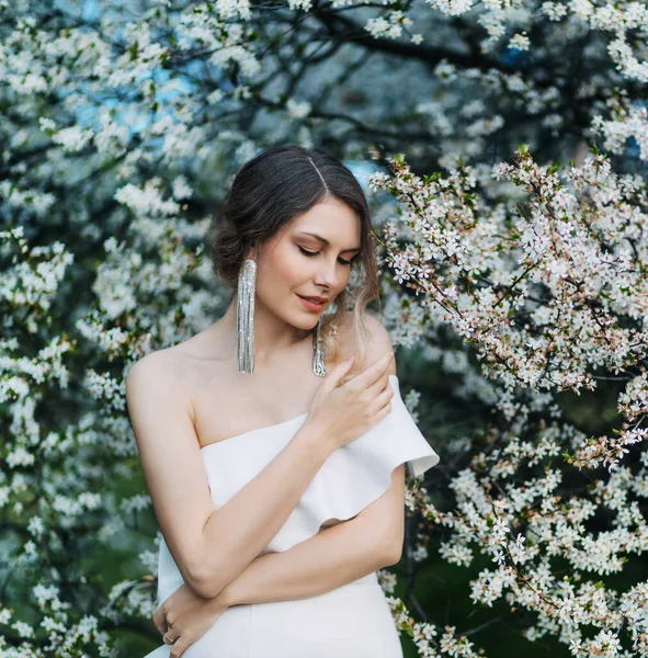 Porträt junge schöne Frau Modell posiert. Hintergrund blühender Baum Frühling Natur weiße Blumen. Elegante Mädchen romantische Frisur gesammelt. lange trendige stylische Ohrringe Schmuckaccessoires — Stockfoto