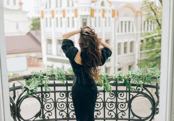 Luxusní žena v černých večerních šatech stojí na balkóně se dívá na město. Zpět na zadní pohled kamery, odvrátil tvář. Záhadná silueta okouzlující dívčí královny. Ruce držet dlouhé vlasy, létat ve větru pohybu — Stock fotografie