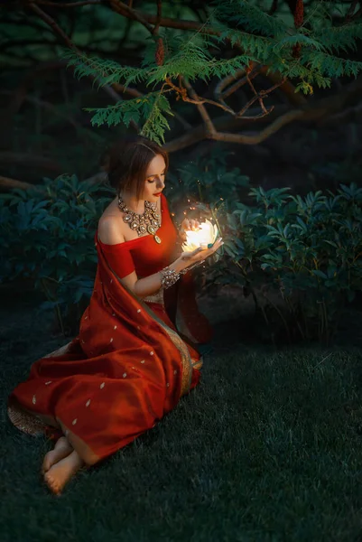 Νεαρή όμορφη ανατολίτικη ινδική φαντασία γυναίκα κάθεται στο πράσινο γρασίδι, φόντο νύχτα μαγικό δάσος. Κόκκινο εκλεκτό φόρεμα σάρι. Κορίτσι κρατώντας μαγικό λαμπερό λουλούδι στα χέρια. Βραχιόλι κόσμημα χρυσό κολιέ — Φωτογραφία Αρχείου