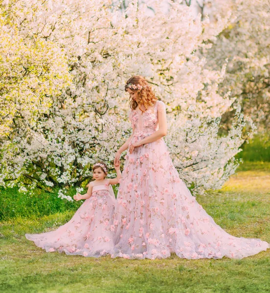 Щаслива красива родина мама і дочка, весняний квітучий сад. Довгі розкішні вечірні фентезійні сукні порошкового рожевого кольору. Королева обережно тримає маленьку принцесу. Зачіска прикрашена квітами — стокове фото