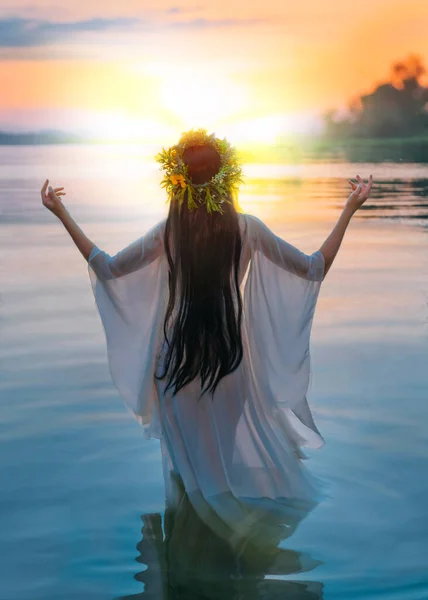 Mulher de fantasia em pé na água mãos levantadas para o céu, orando ao sol. Menina eslovena em grinalda de ervas na cabeça, vestido branco de cabelo longo, férias pagãs de Ivan Kupala. Pôr-do-sol azul natureza. Visão traseira. — Fotografia de Stock