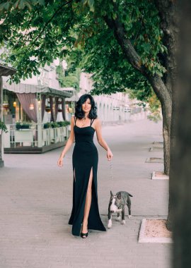 Mutlu orta yaşlı kadın teriyer cinsinden sevgili bir köpekle yürüyor. Cafe 'nin arka planı, açık sokak restoranı. Esmer, dalgalı saçlı, siyah akşam tarzı, seksi, uzun elbiseli bir kadın. Gülen surat.