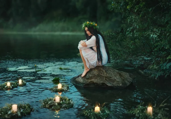 Słowianka siedzi na kamieniu nad brzegiem jeziora. Nimfowa kobieta fantazji przytula kolana. Długie czarne włosy. Wieńce trawy, kwiaty unoszą się na wodzie. Świece płoną. Rzeka zmierzch las zielony drzewo. Rytuał Wróżbiarstwa — Zdjęcie stockowe