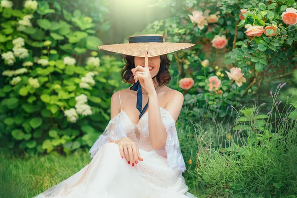 Lekfull glad glad fantasi kvinna döljer sitt leende ansikte under bred brättad båtägare hatt Lady sitter på grönt gräs. Vit vintage bröllop klänning boho stil. Sommar natur träd blommar rosa blommor — Stockfoto