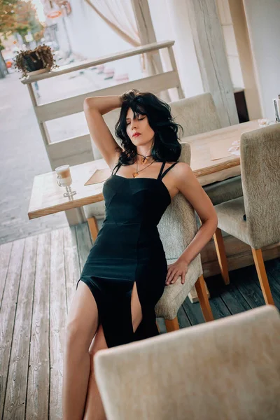 Donna fantasia sexy si trova sulla sedia caffè strada. Lady divertirsi sulla terrazza ristorante della città. Ragazza bruna tocca delicatamente i capelli ondulati con la mano. Abito lungo romantico sexy nero. Panorama urbano sullo sfondo — Foto Stock