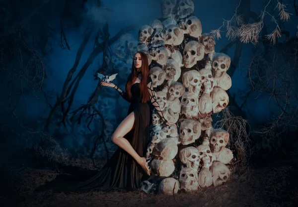 Fantasy gothic redhaired woman dark queen stands near mountain of skulls, bones. Lady goddess of death in black long creative dress, holds dragon on hand. Vampire girl princess. Night forest tree, fog — Stock Photo, Image