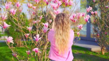 Mutlu portre güzel kadın elleri çiçek açan ağaçlara dokunuyor. Pembe manolya çiçeği dalı. Yetişkin kız, çiçek aromasının tadını çıkarıyor. Bahar yeşili güneş ışığı. sarı saçlar.